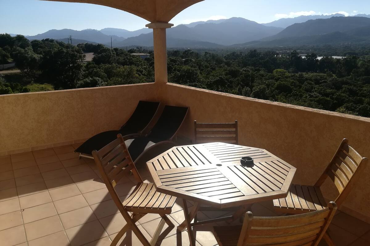 A balcony with a wooden table and chairs, offering a view of distant mountains and greenery.