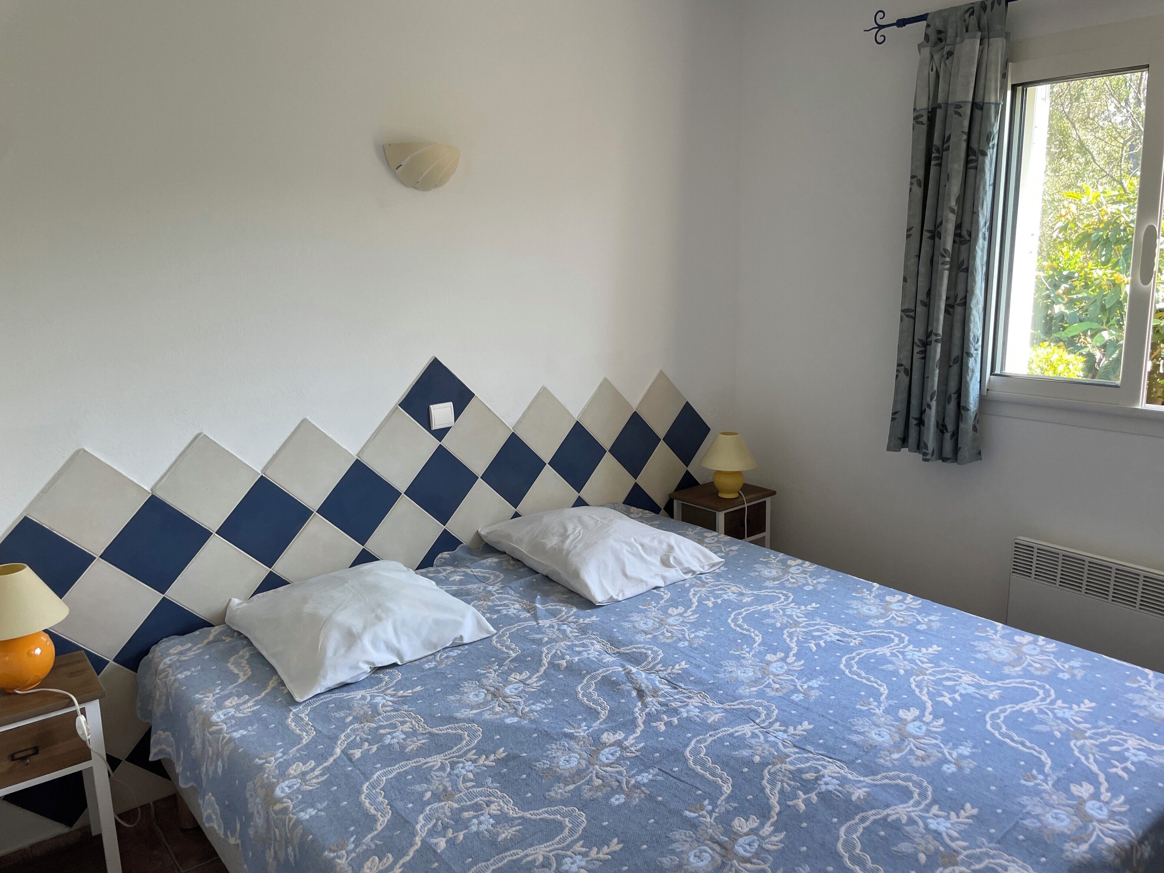 A bedroom with a bed, a nightstand, a lamp, a wall-mounted light, and a window with curtains.