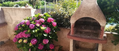 A stone-built outdoor oven, a flower bed with pink flowers, and a metal fence in a garden setting.