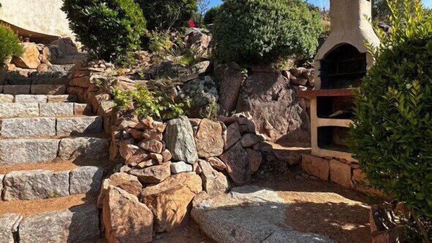 Una escalera de piedra que conduce a un edificio con una chimenea, rodeado de vegetación y rocas.