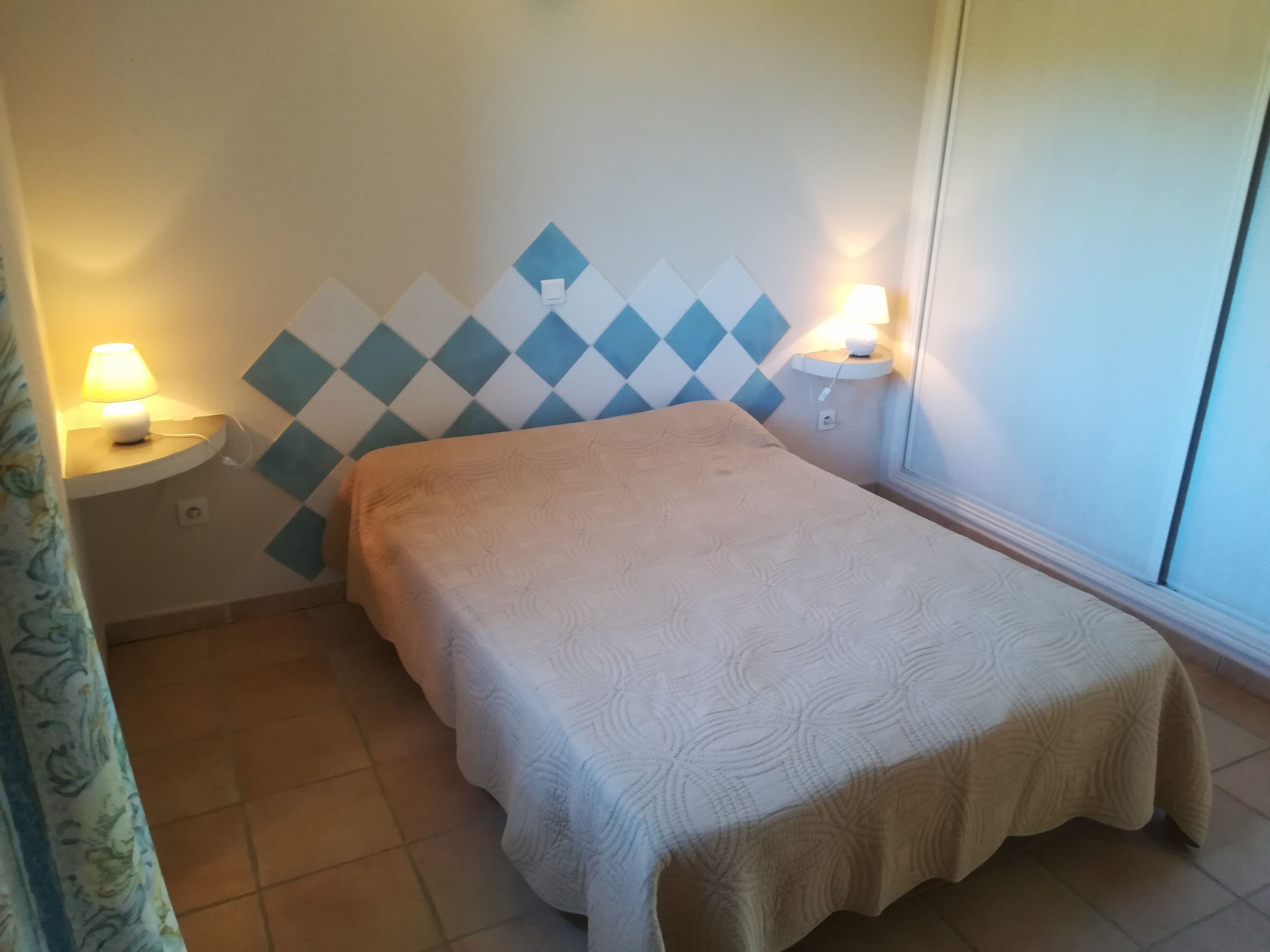 A small, neatly made bed with a patterned bedspread in a room with a tiled floor and a wall decorated with blue and white diamond patterns. There are two bedside lamps on white nightstands.