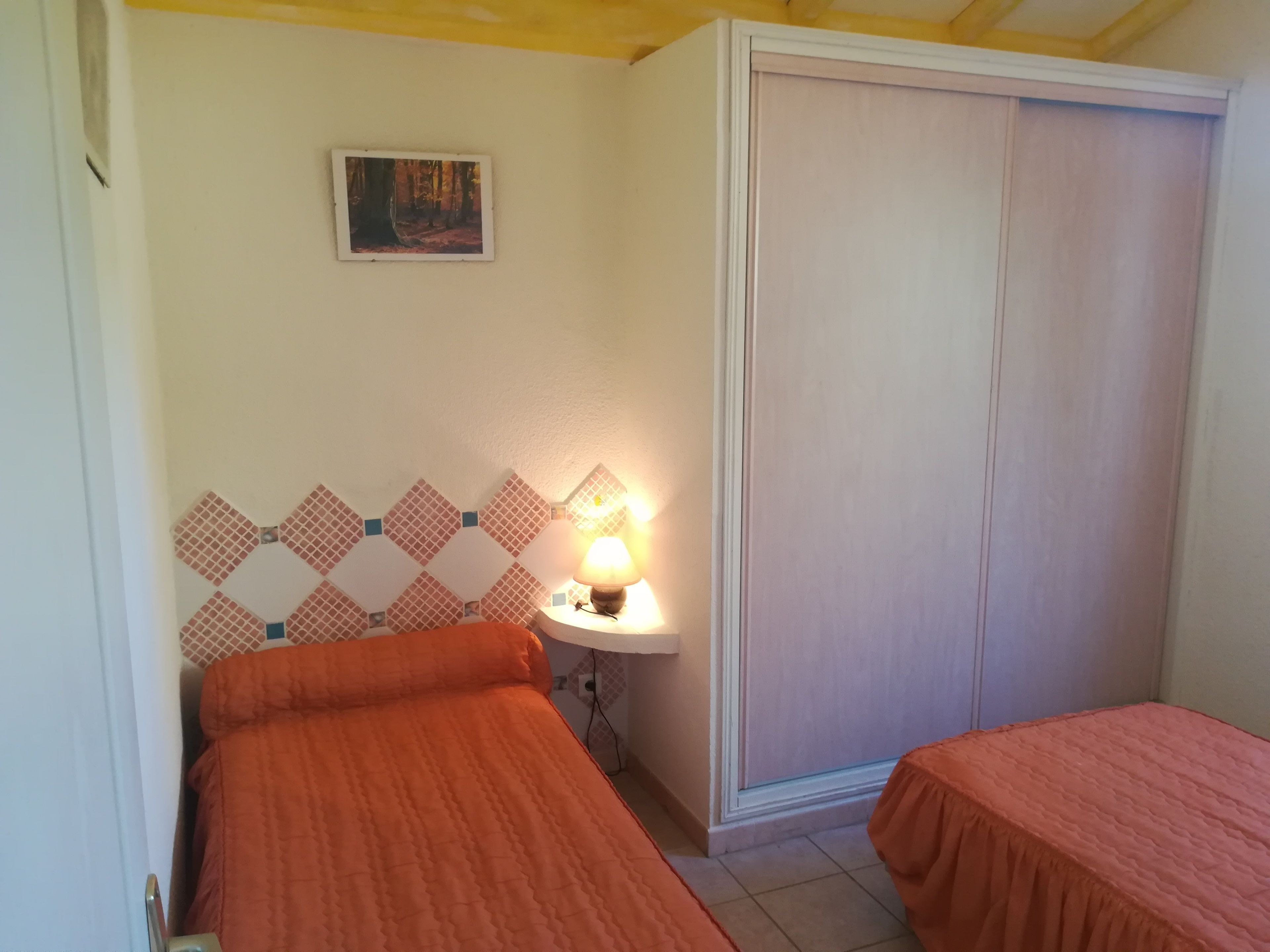 A bedroom with two beds, a nightstand with a lamp, a wall-mounted picture, and a wardrobe.
