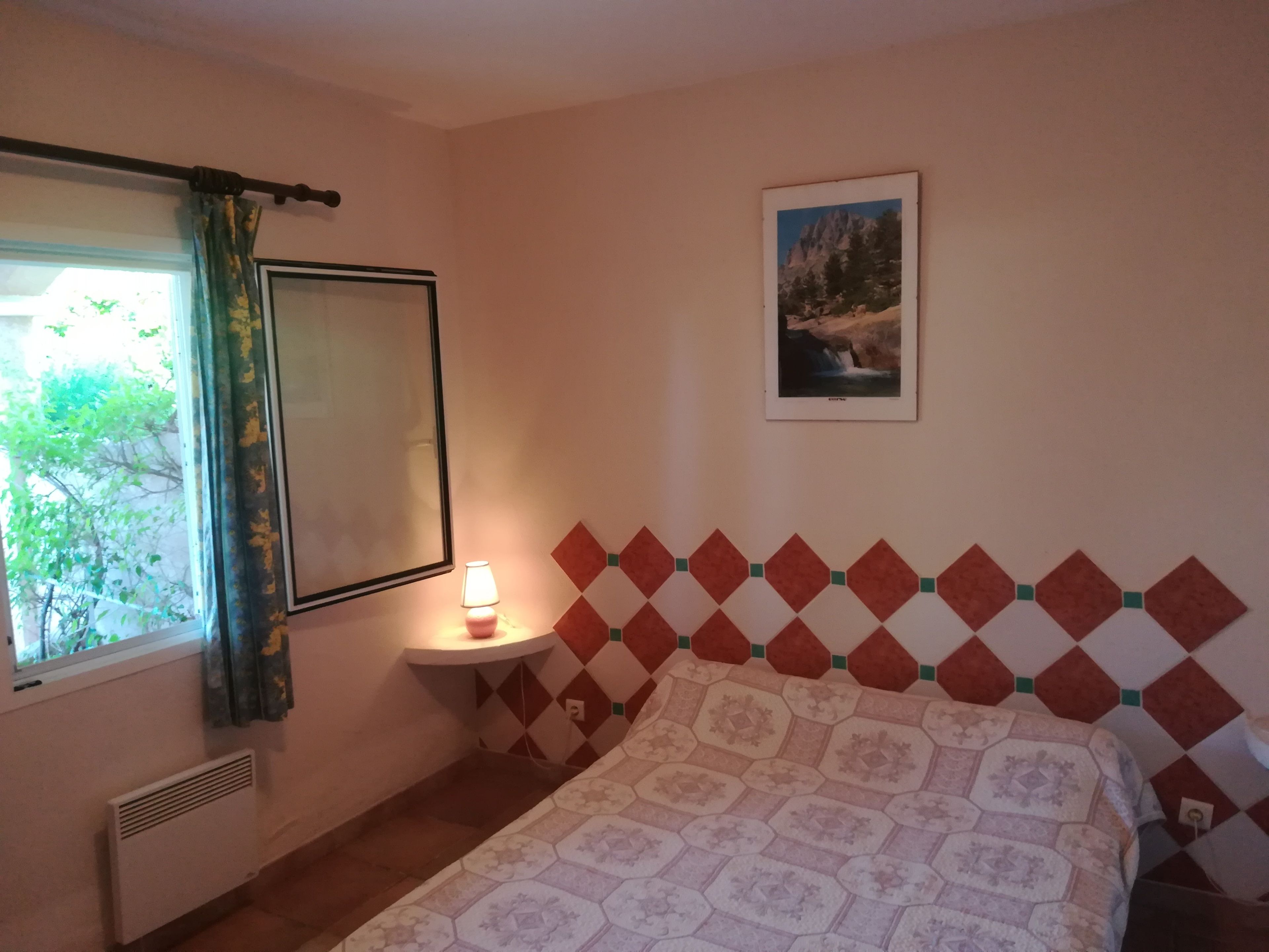 A bedroom with a bed, a bedside table with a lamp, a window with curtains, a radiator, and a wall-mounted picture.