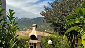 Un espacio al aire libre rústico con un sendero de piedra que lleva a un horno de piedra tradicional, rodeado de exuberante vegetación y una vista despejada de montañas distantes.