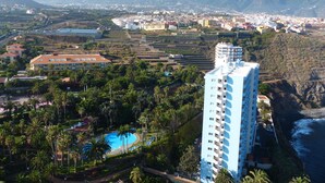 A coastal hotel with a pool, surrounded by palm trees and a mountainous backdrop.