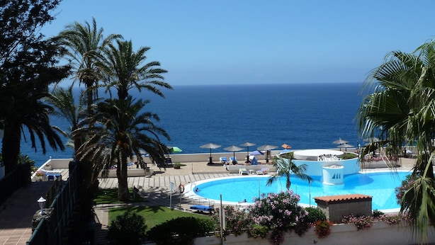 A resort pool with a view of the ocean, surrounded by palm trees and lounge chairs.