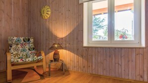 Una habitación con paneles de madera, una ventana que da a zonas verdes, una silla con estampado floral y una lámpara sobre una mesita.