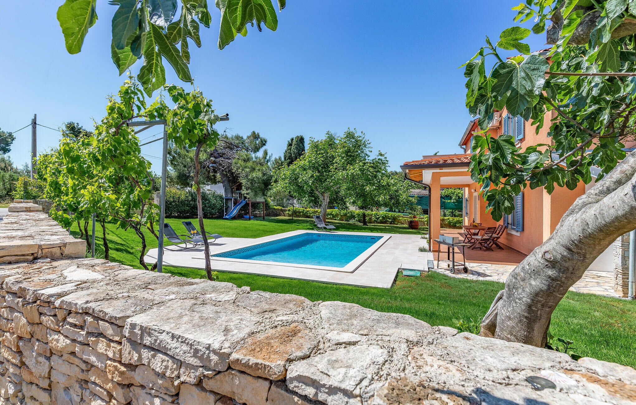 A swimming pool with a stone wall in the foreground, a grassy lawn, and a building with a tiled roof in the background.