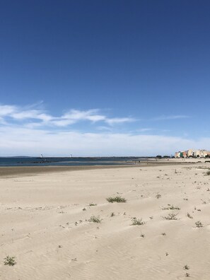Ein weiter Sandstrand mit vereinzelter Vegetation, ein klarer blauer Himmel und Gebäude in der Ferne.