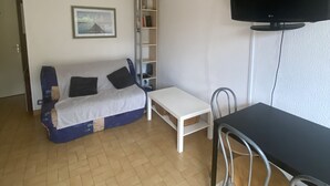 A small, furnished room with a sofa, a white table, a TV, and a bookshelf.