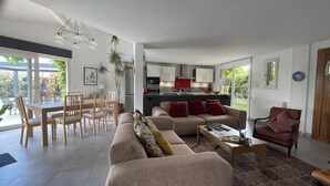 Un salon moderne avec un canapé, une table basse et un coin repas. Au fond, on aperçoit une cuisine avec des meubles blancs et un plan de travail rouge.