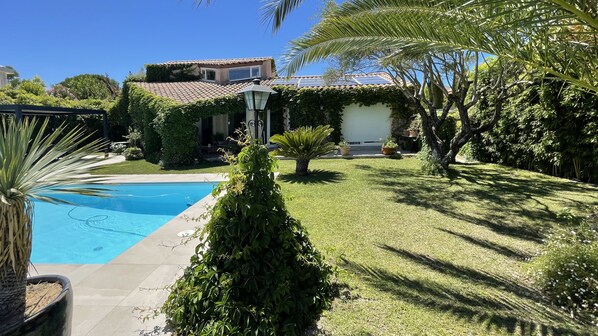 Una piscina rodeada de exuberante vegetación, una casa con techo de tejas y un césped bien cuidado.
