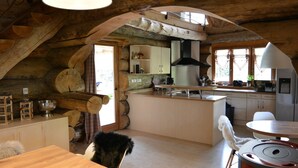 A rustic cabin interior with a wooden dining table, a fur-covered chair, a kitchen area with a stove, and a large black pendant light. - 5-star log cabin with SPA (Erckartswiller)
