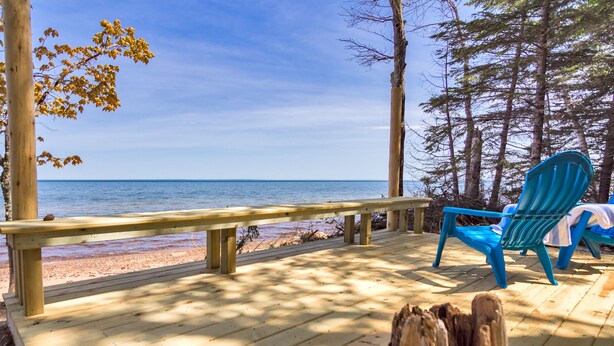 A wooden deck with a bench and two blue Adirondack chairs overlooking a beach.