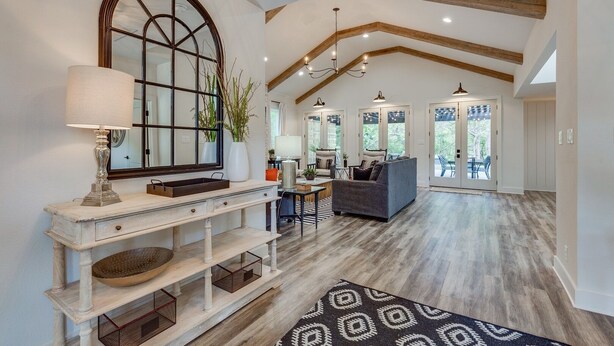 A spacious living area with a high ceiling, exposed wooden beams, a large window with a mirror, and a modern chandelier.
