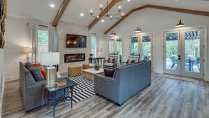A spacious living room with a fireplace, a large TV, and a dining area with outdoor seating visible through glass doors.