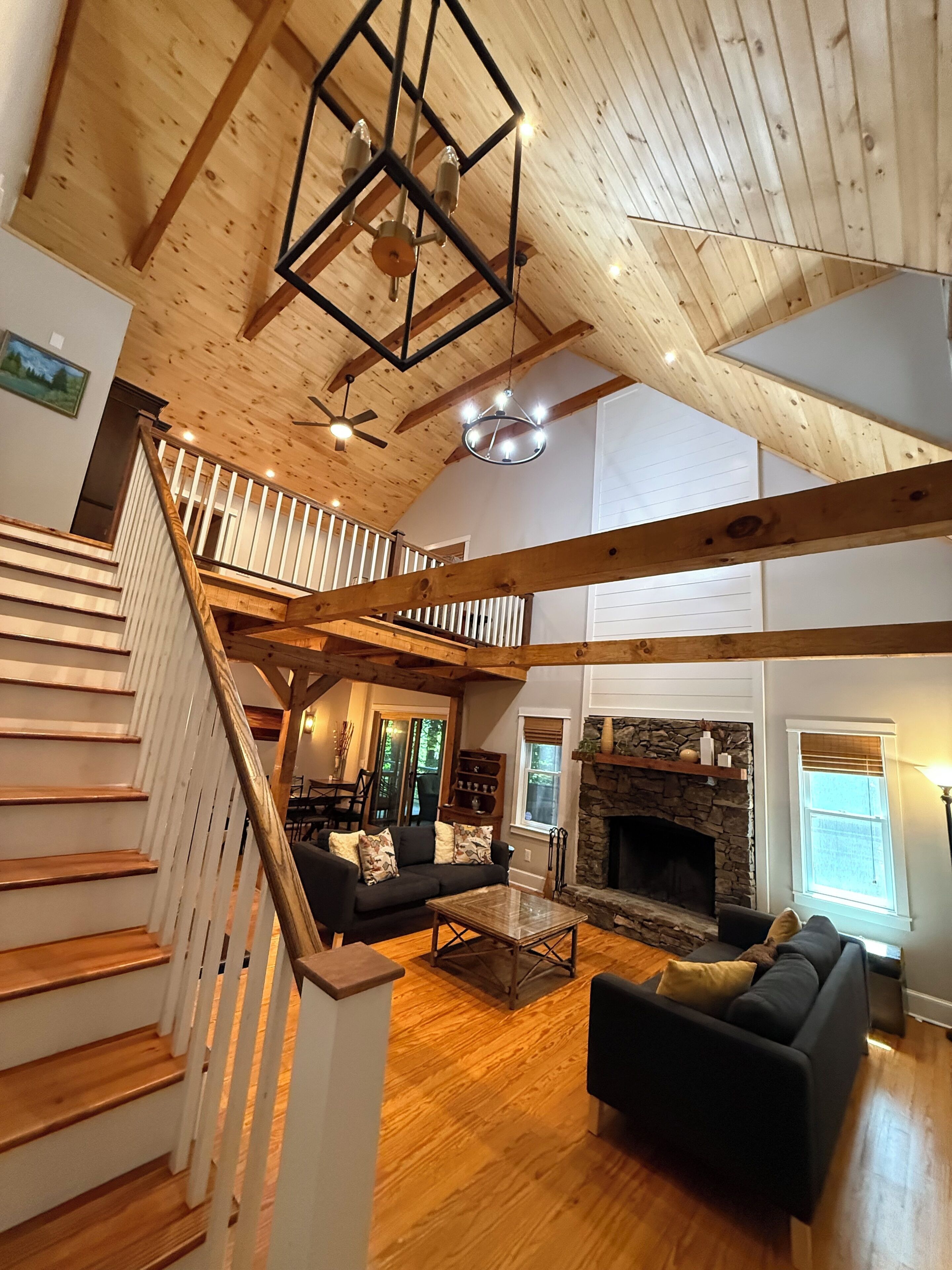 A spacious living area with a wooden staircase, a stone fireplace, and a high ceiling with exposed beams.