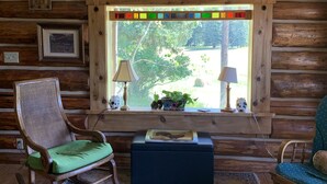 L’intérieur chaleureux d’un chalet avec un mur en rondins de bois, un fauteuil à bascule doté d’un coussin vert, une petite table avec une lampe et une fenêtre ornée d’un vitrail aux couleurs vives.