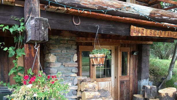 Une cabane rustique avec un mur en pierre, des poutres en bois et une enseigne indiquant « LAZY BEAN RANCH ».