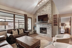 A spacious living room with a stone fireplace, leather sofa, and a view of a cityscape through large windows. - Ski In Ski Out 2 Bed 3 bath, 1100sqft. Vaulted ceiling sleeps 10 in Ctr Village (Frisco)