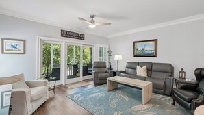 A living room with a ceiling fan, a sign reading 'THE GATHERING PLACE,' a large window, and a view of greenery outside. - Award-Winning Resort Amenities | Grand Pavilion 10 (Isle of Palms)