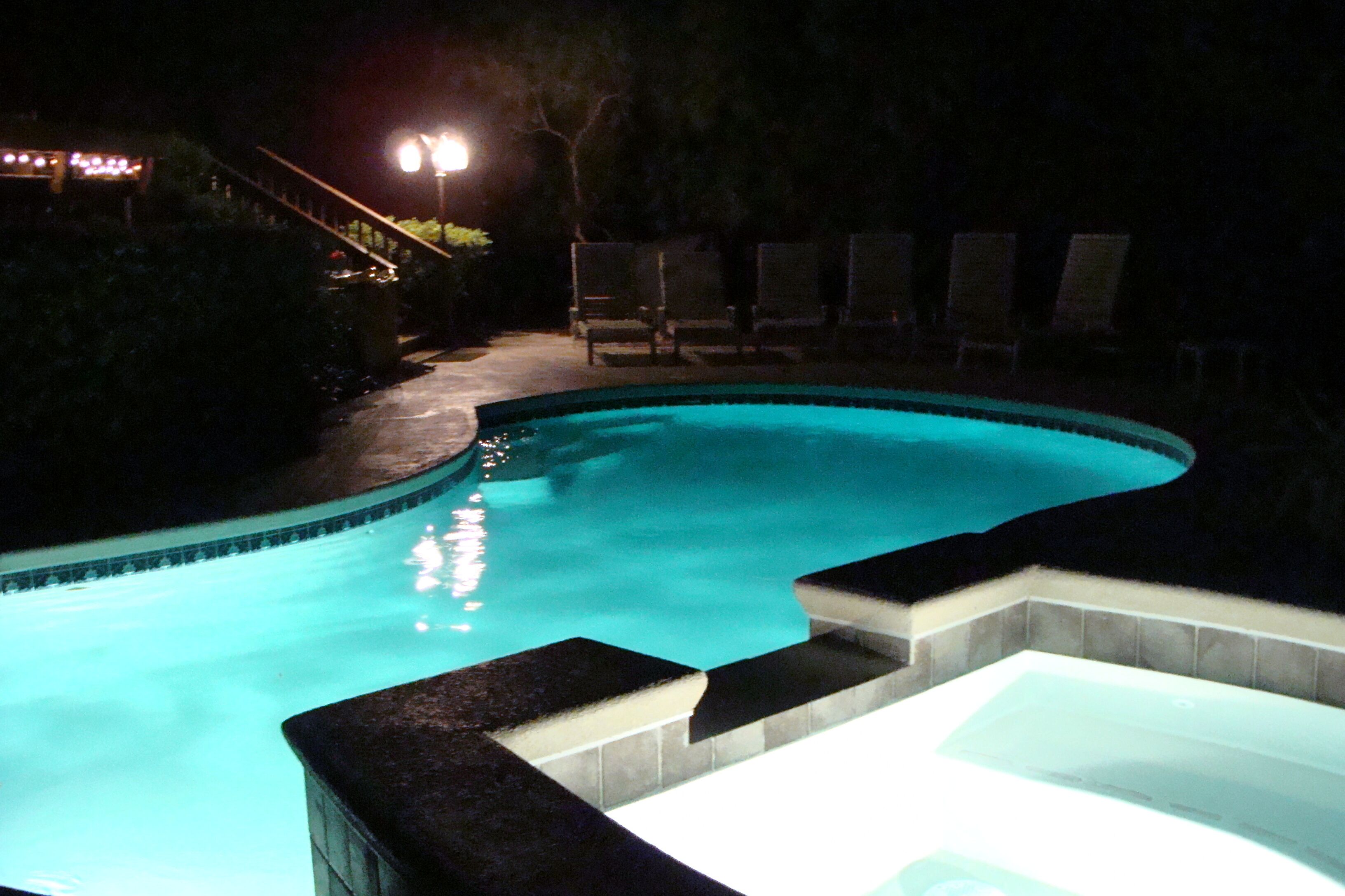 Una piscina nocturna con un jacuzzi, rodeada de sillones de descanso y una escalera que conduce a una terraza.