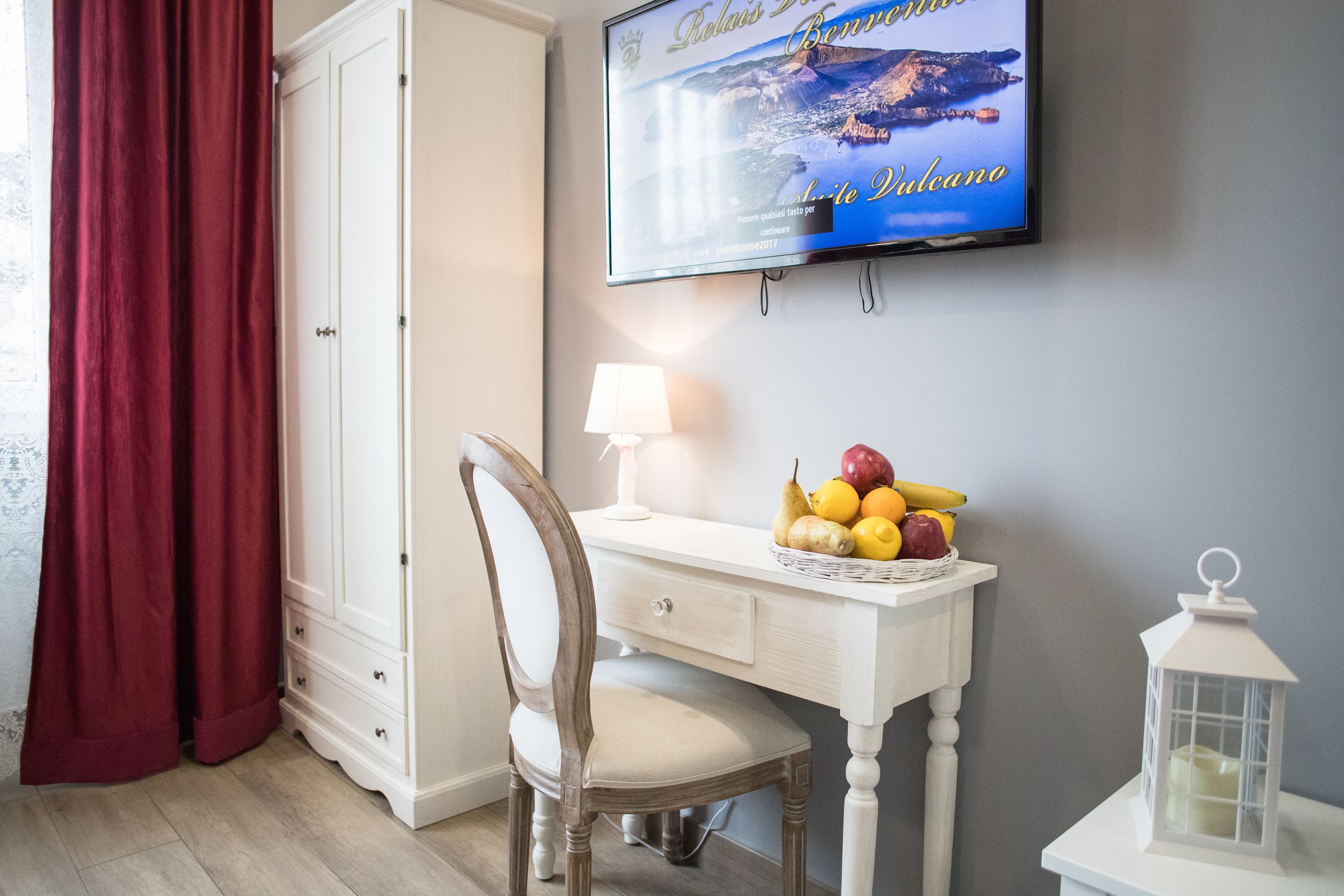 A room with a TV displaying a travel show, a white desk with a lamp and fruit basket, a wooden chair, and a red curtain.