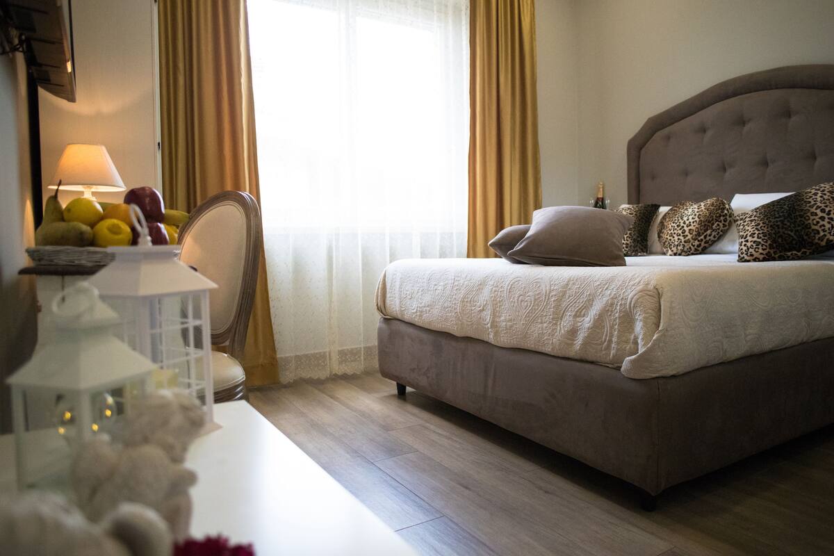 A neatly arranged bedroom with a large bed, a bedside table with a lamp, and a window with curtains.