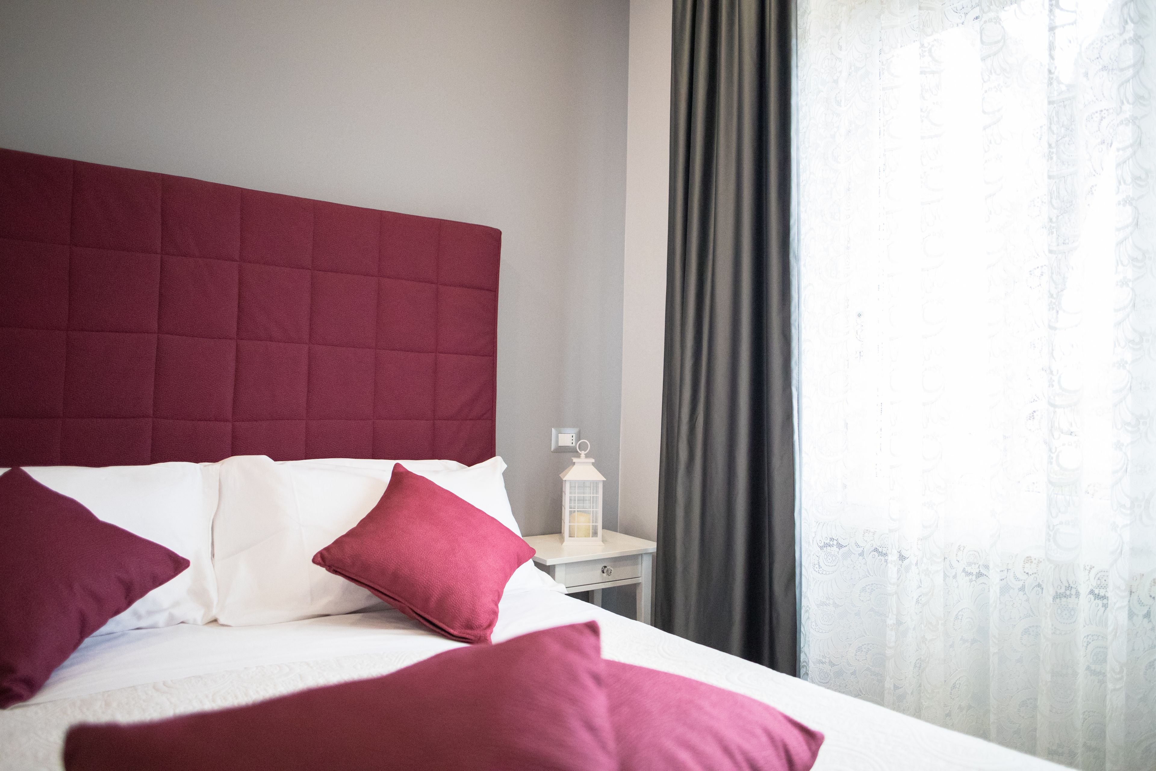 A neatly made bed with a red headboard, white bedding, and matching red pillows. A small bedside table with a white lantern is visible.