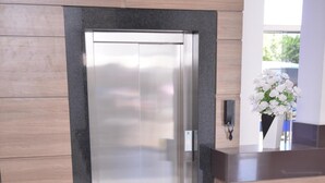 Un área de recepción moderna con un ascensor de vidrio, paneles de madera y suelo de baldosas blancas.