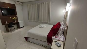 Habitación de hotel con cama, escritorio y silla, refrigerador pequeño, televisor montado en la pared y ventana con cortinas.