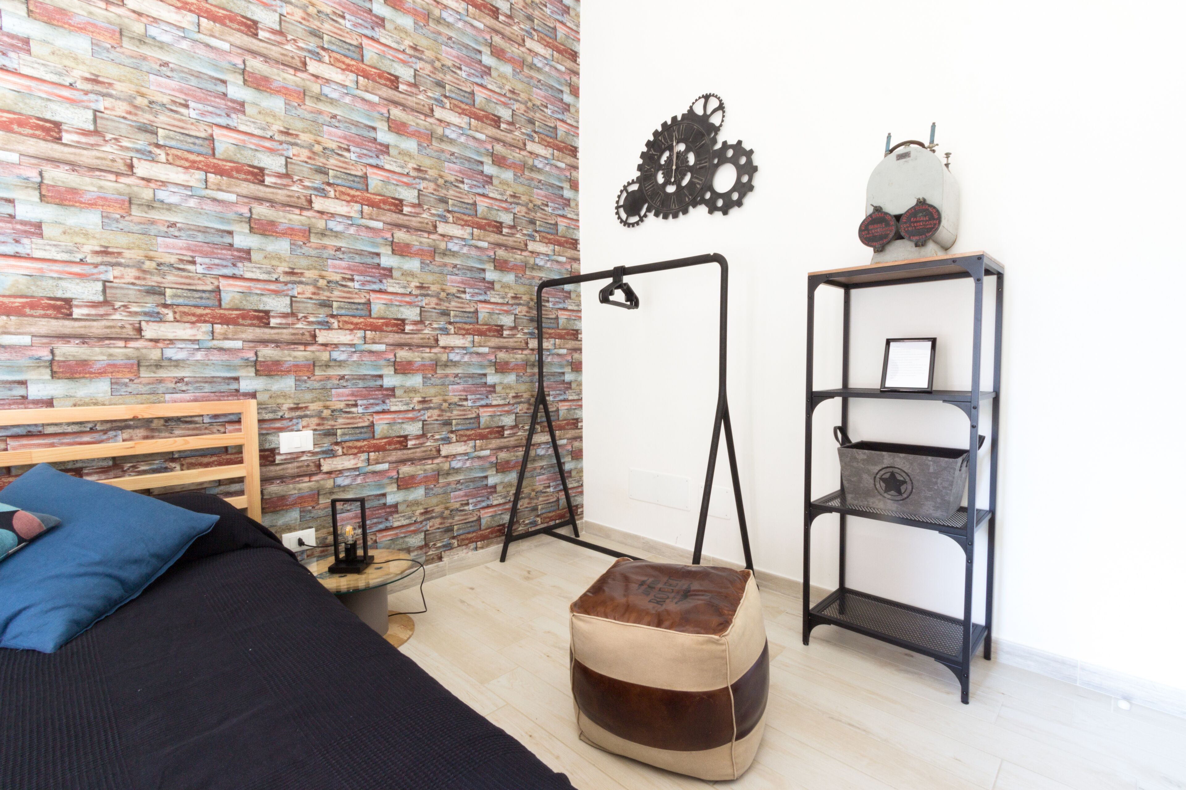 Una habitación con pared de ladrillo, una cama con almohadas azules, una mesita con una lámpara, un taburete y una estantería metálica con objetos decorativos.