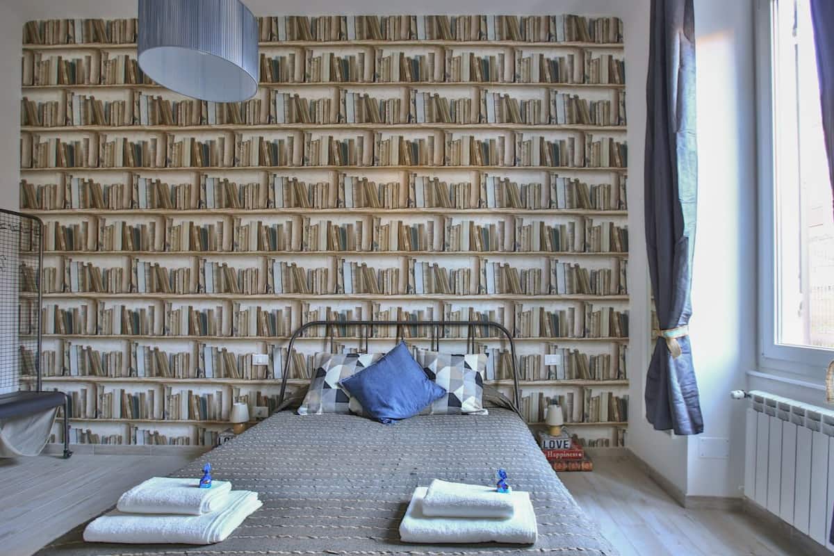 A bedroom with a large bookshelf wall, a bed with two towels, and a window with curtains.