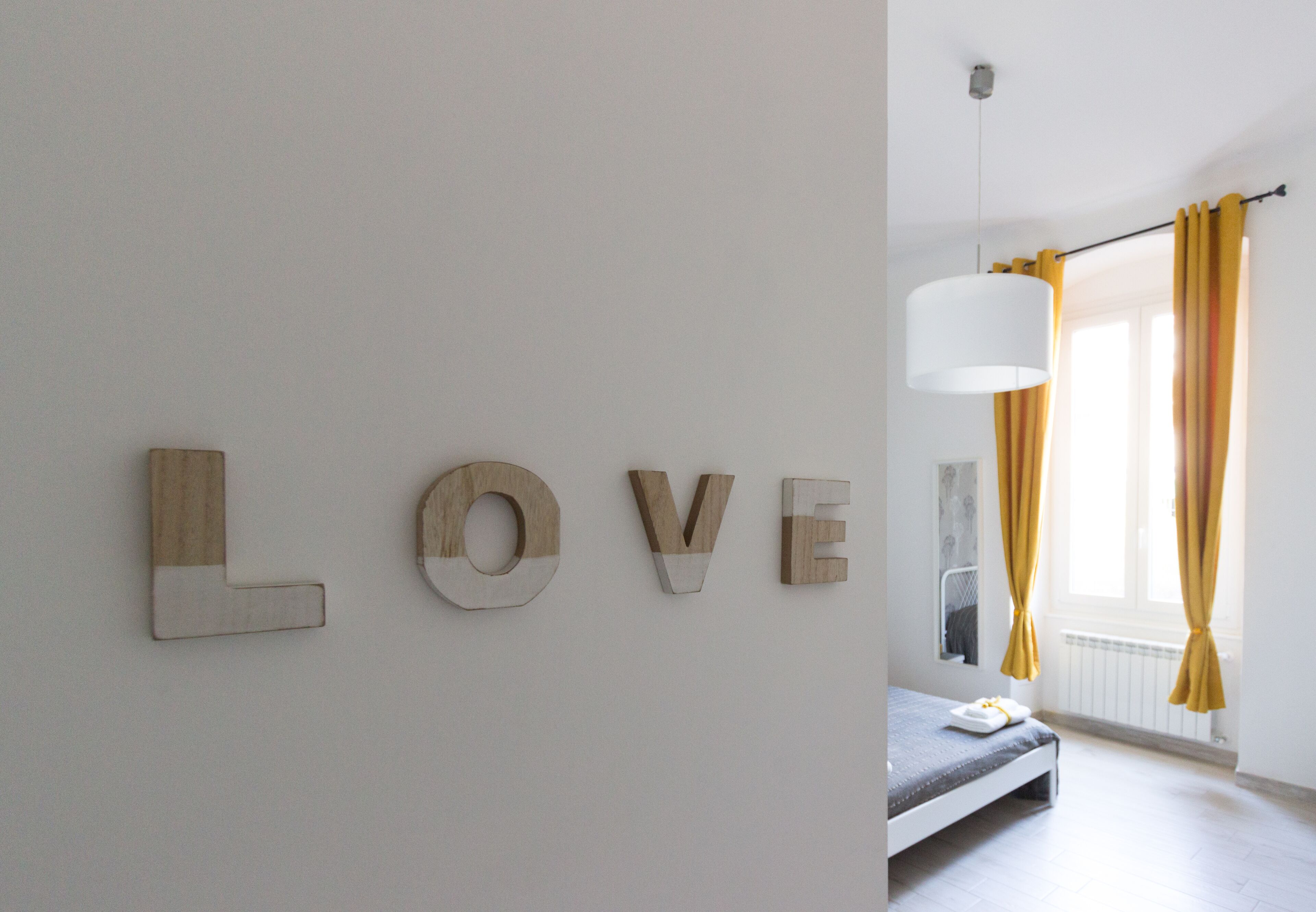 Une chambre à coucher avec un lit, un mur sur lequel le mot « AMOUR » est écrit en lettres de bois, et une fenêtre avec des rideaux jaunes.