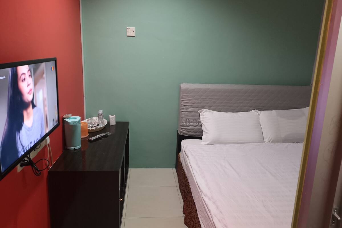 Una habitación de hotel pequeña con cama, televisión, cómoda y enchufe de pared.