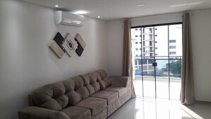 Een moderne woonkamer met een beige bank, een groot raam met gordijnen en uitzicht op een gebouw.