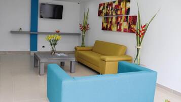 Una sala moderna con un sofá amarillo, un sillón azul y una mesa de centro gris. A cada lado del sofá hay dos jarrones grandes con plantas.