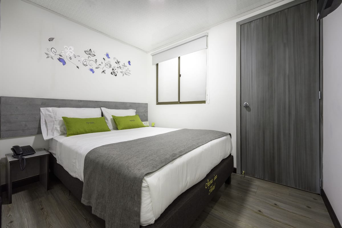 A neatly made bed with a grey and white duvet, a green pillow, and a grey throw blanket. There is a bedside table with a phone and a lamp. The room has a wooden floor and a grey wardrobe.
