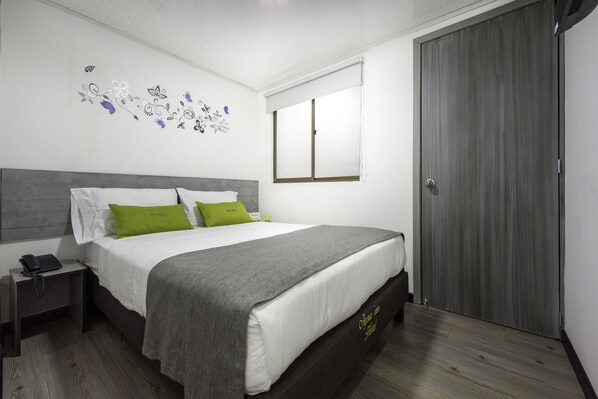 A neatly made bed with a grey and white duvet, a green pillow, and a grey throw blanket. There is a bedside table with a phone and a lamp. The room has a wooden floor and a grey wardrobe.