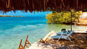 Vista de la playa con aguas azules cristalinas, una estructura de techo de paja y dos sillas de lounge blancas.