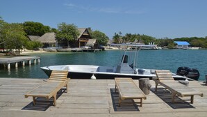Un muelle de madera con sillones de descanso, un bote y una construcción con techo de paja junto al agua.