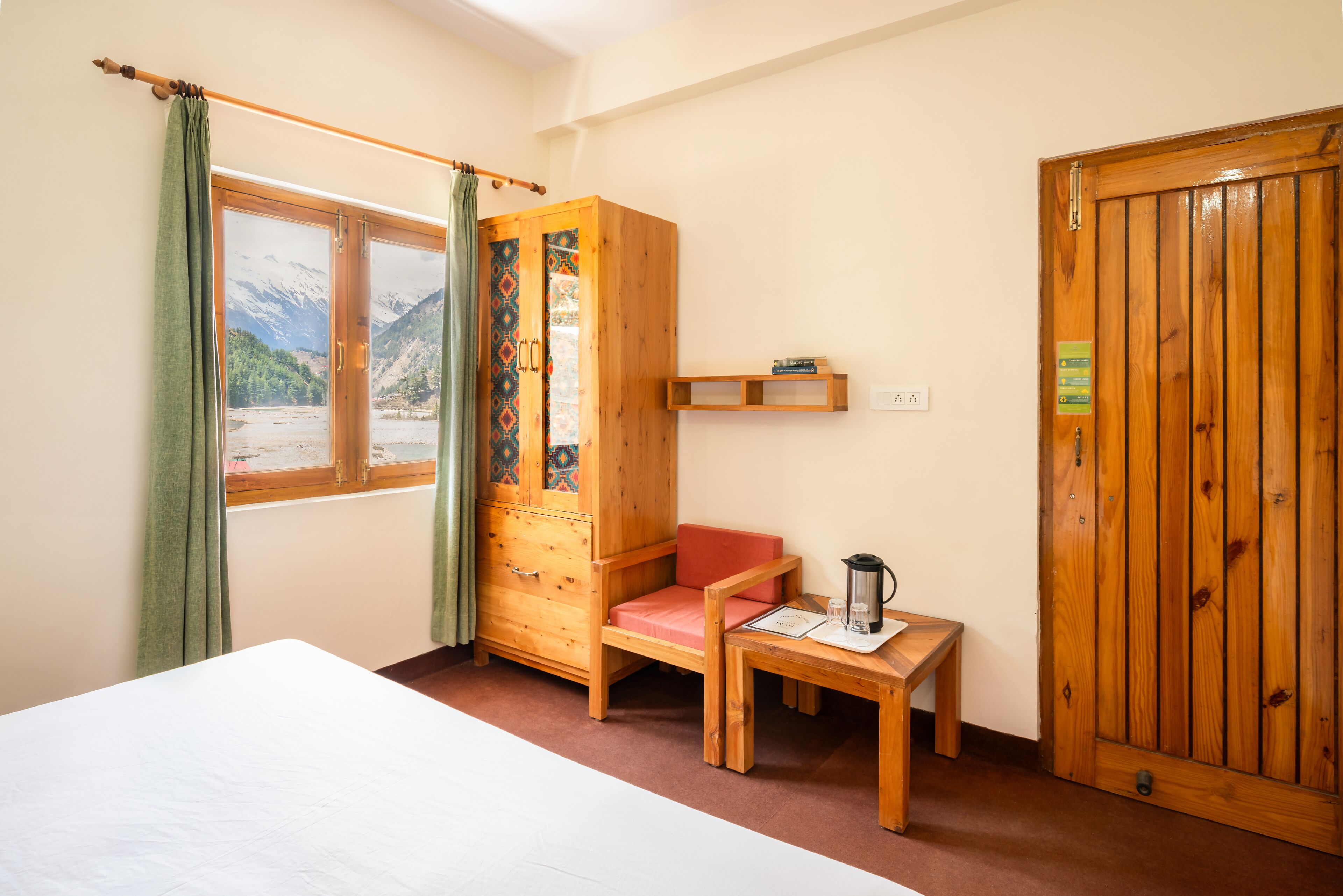 A room with a bed, a wooden wardrobe, a small table with a kettle, and a window with a view of mountains.