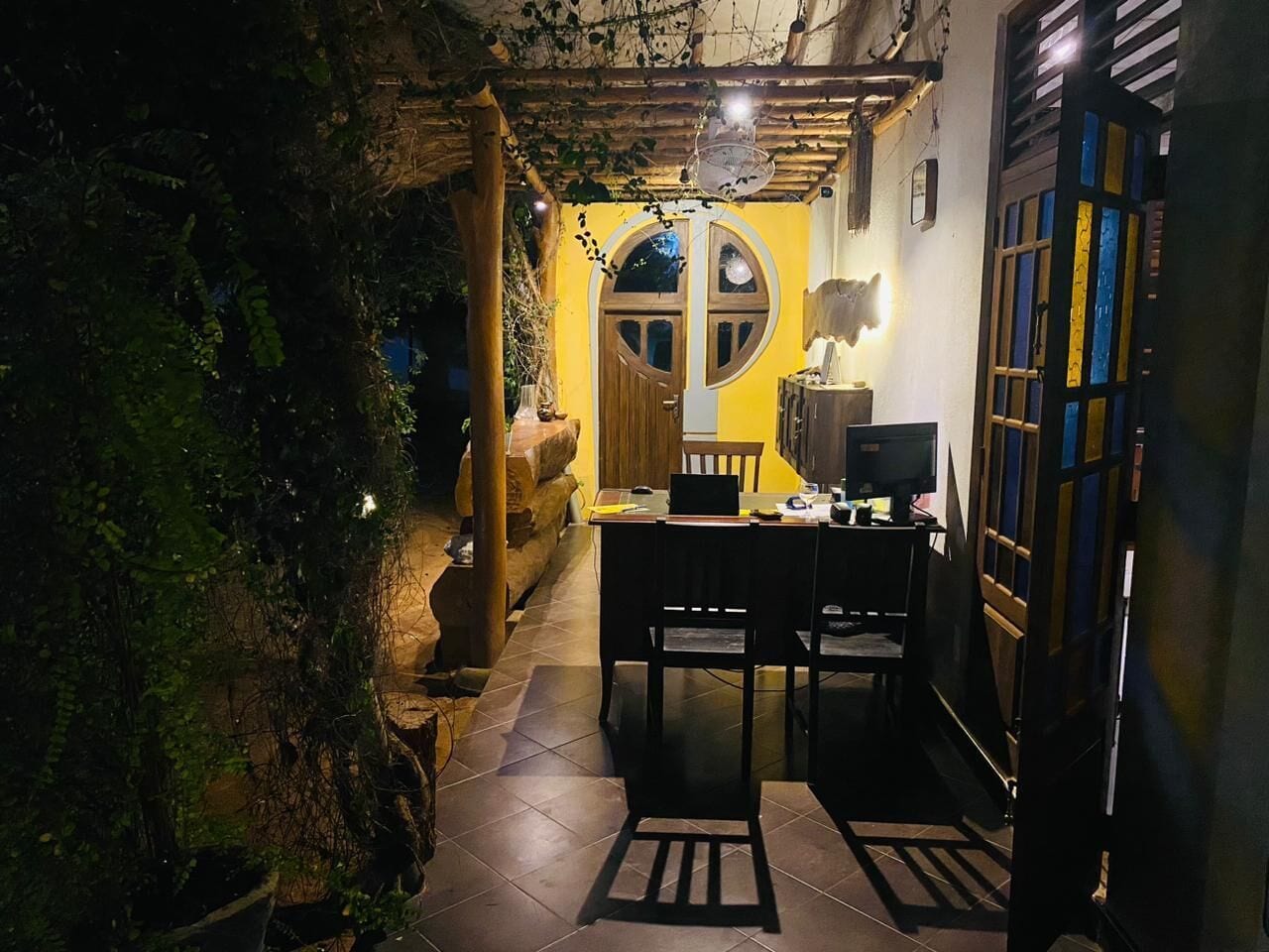 A narrow outdoor corridor with a table and chairs, a wooden door with a circular window, and a wall with a yellow hue.