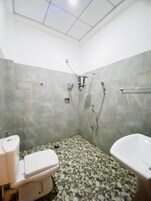 Ein Badezimmer mit Toilette, Waschbecken und Badewanne. Der Boden ist mit Kieselsteinen belegt, die Wände sind mit Fliesen versehen.