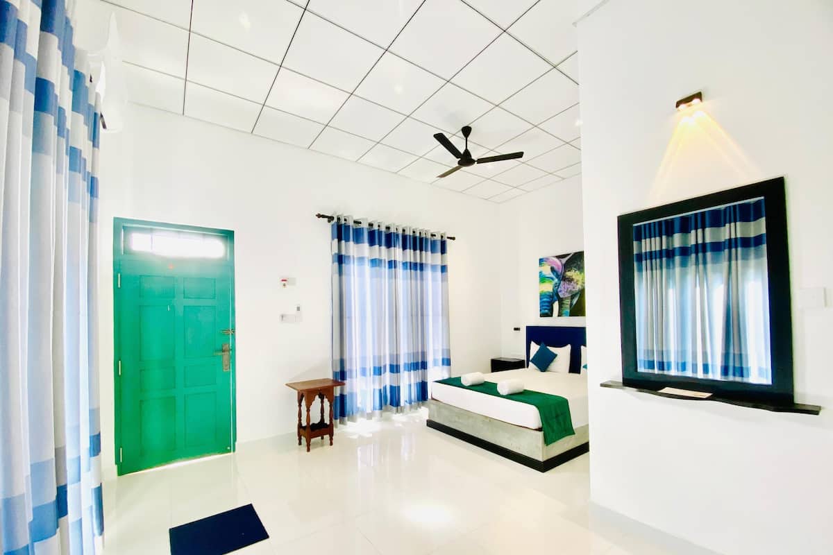 Habitación de hotel con puerta verde, cama con cubrecama verde, ventilador de techo y ventana con cortinas azules.