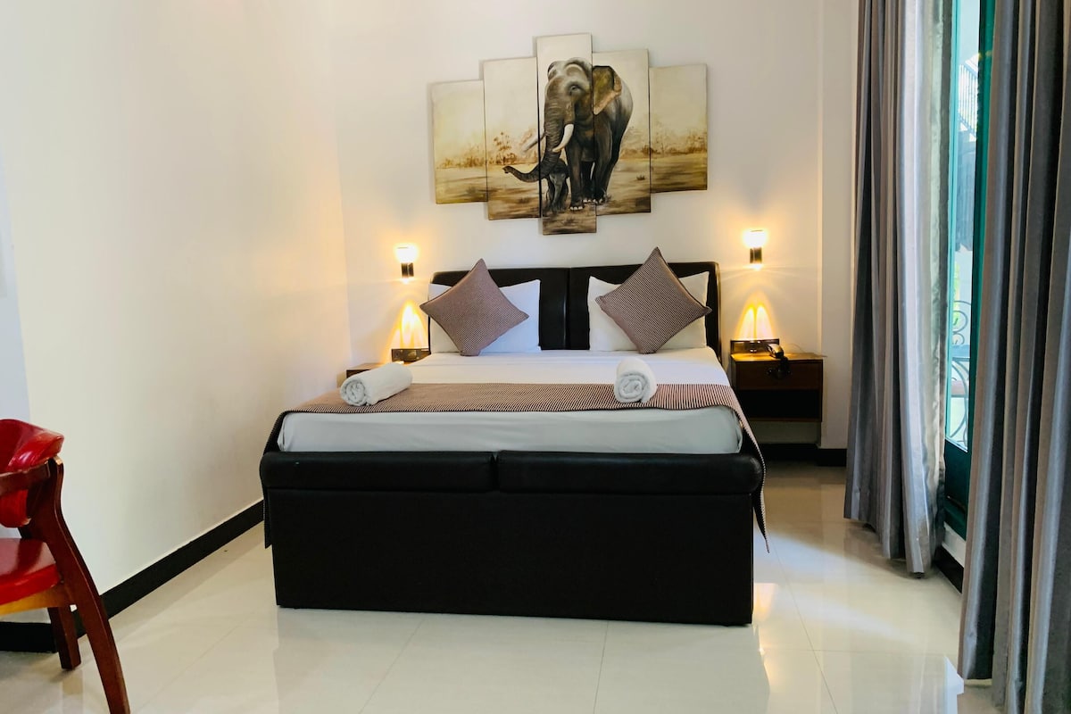 Habitación de hotel con una cama grande, dos mesitas de noche con lámparas, una silla roja y un cuadro de elefantes en la pared.