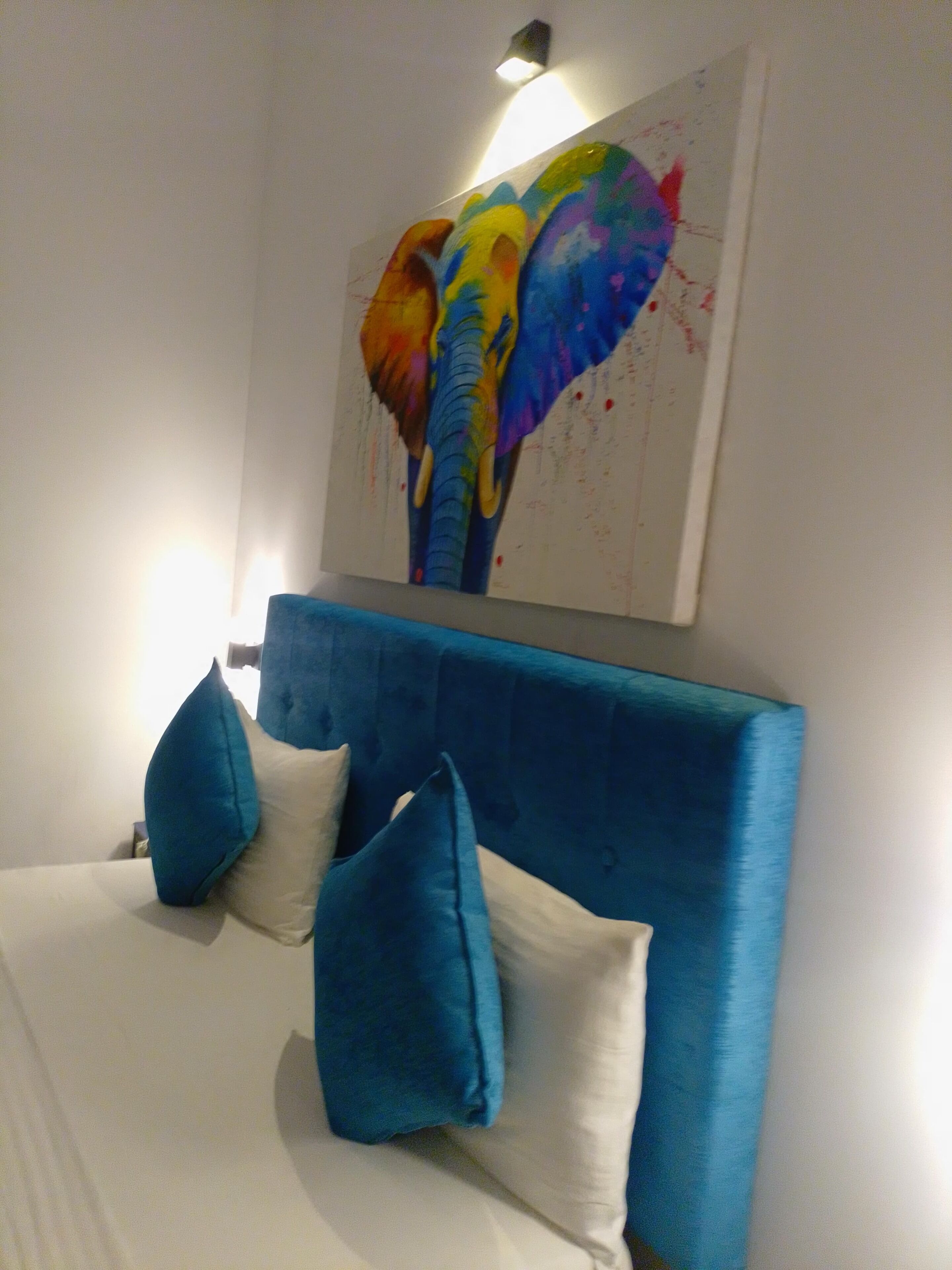 A bed with a blue headboard and two pillows, a painting of an elephant on the wall, and a light fixture above the painting.