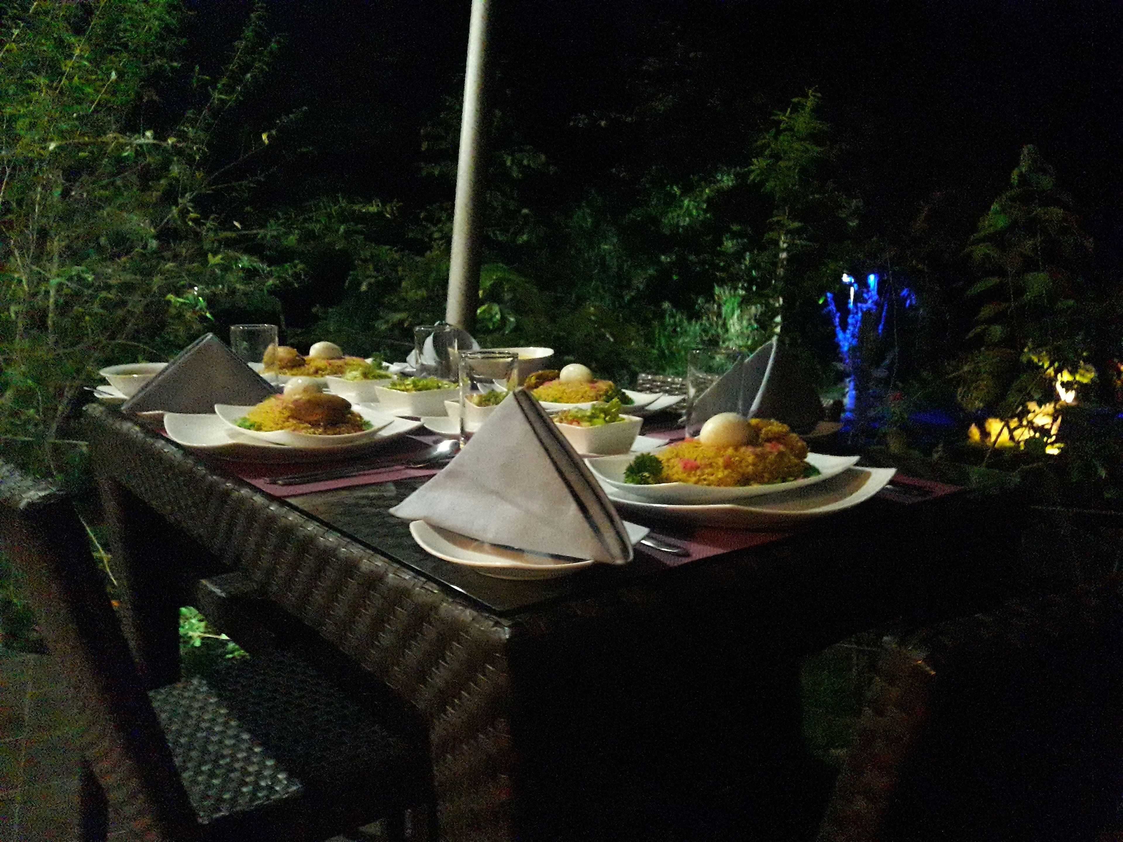 Mesa de refeições ao ar livre posta com pratos de comida, copos e talheres, rodeada por cadeiras e uma luminária decorativa.