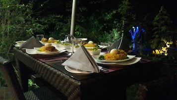 Mesa de refeições ao ar livre posta com pratos de comida, copos e talheres, rodeada por cadeiras e uma luminária decorativa.