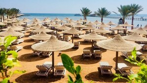 Una playa con filas de tumbonas y sombrillas de paja, palmeras y un muelle que se adentra en el mar.